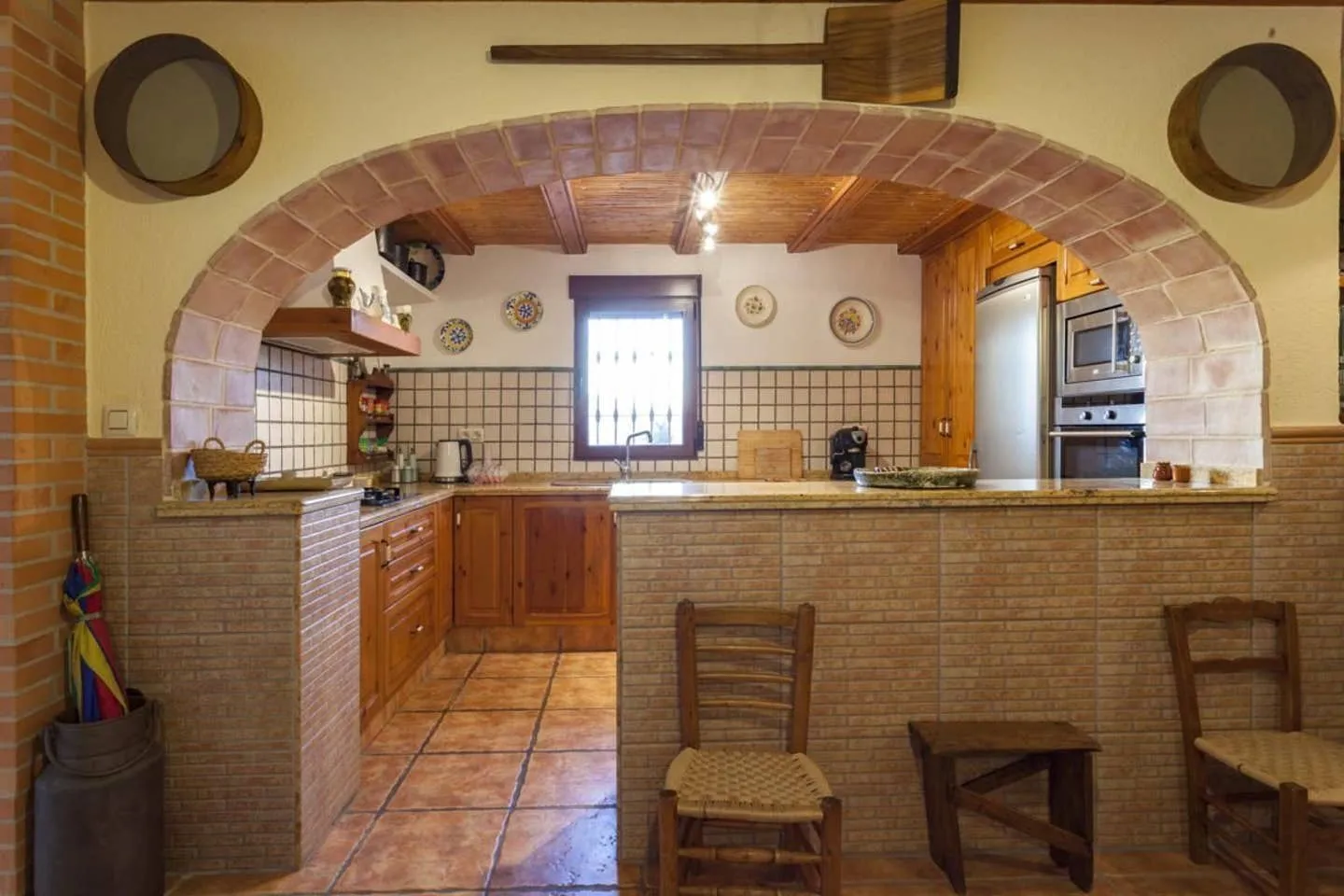 Cocina con barra americana y taburetes de anea, enmarcada por un arco de ladrillo visto, con horno, microondas y nevera