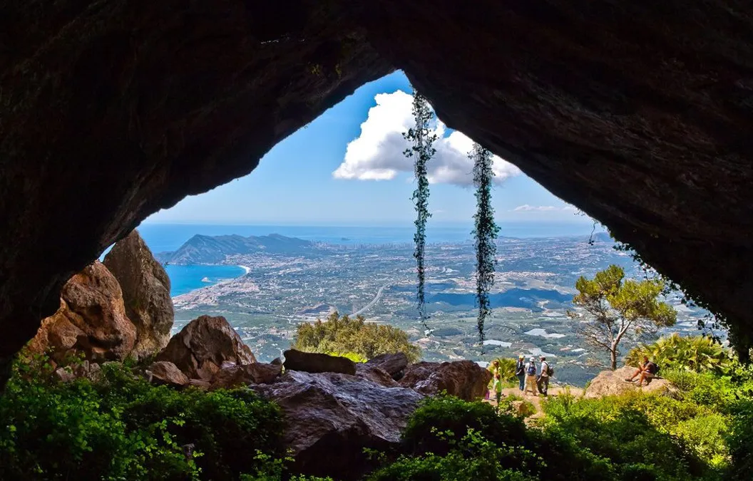Vista desde la boca de una cueva de la sierra de Bèrnia: excursionistas en el sendero, pinos y, al fondo, la costa y el mar Mediterráneo